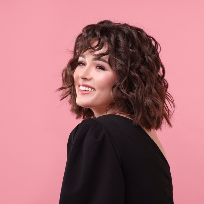 Coiffure Cheveux Bouclés : Femme Brune Souriante sur Fond Rose Femme brune souriante avec une coupe au carré bouclée et une frange sur fond rose uni.
